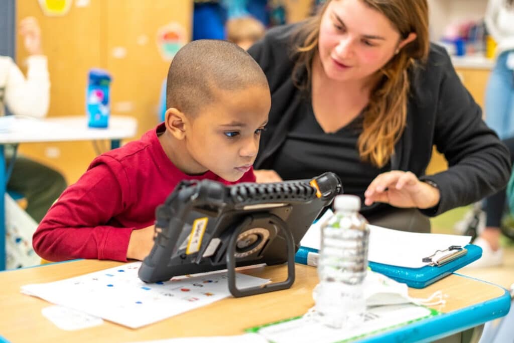 A boy uses a QuickTalker Freestyle speech device at his classroom desk while a teacher leans in beside him