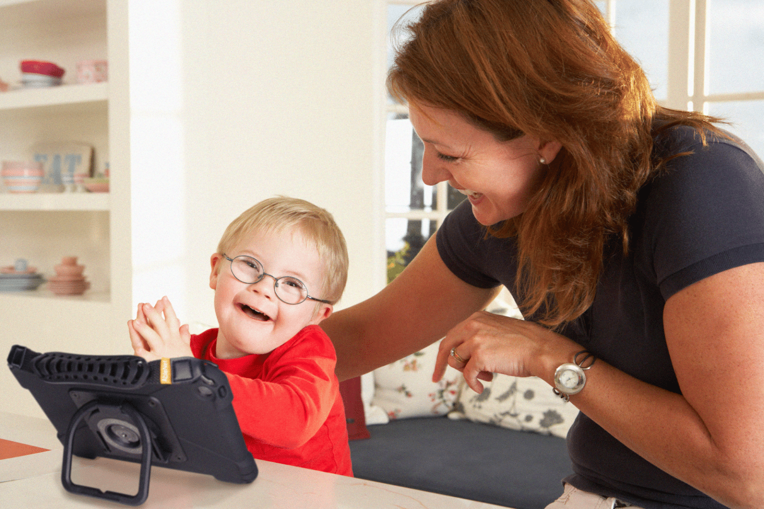 Child joyfully learning with a tablet alongside a smiling caregiver in a bright setting.
