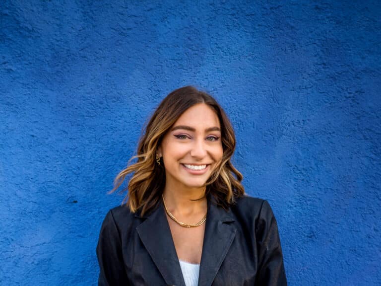 Smiling woman in front of a vibrant blue wall, showcasing contemporary fashion and joy.