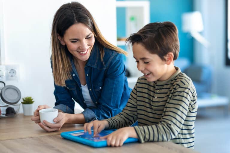 Mother enjoys coffee while her son learns on a digital tablet at home.