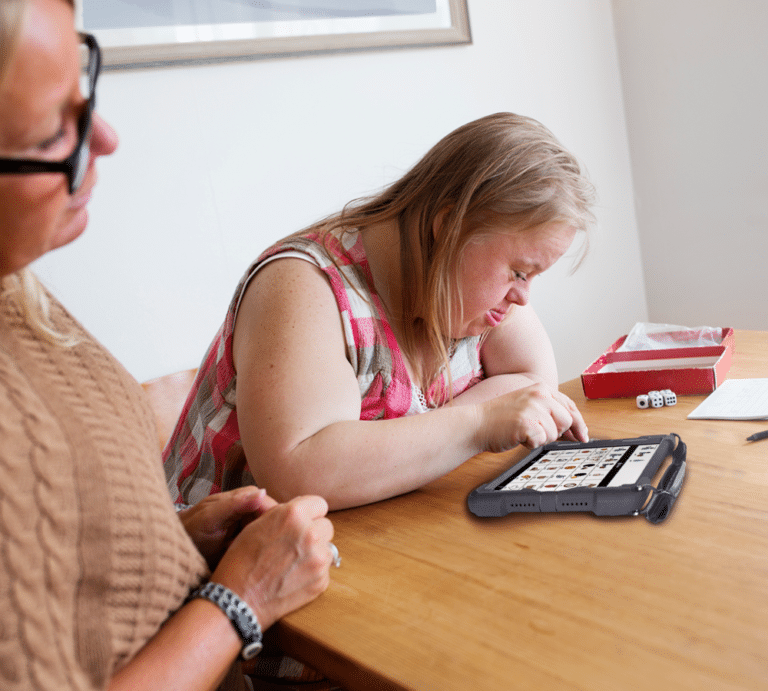 Two women enjoy playing bingo on a high-tech AAC device at a wooden table.