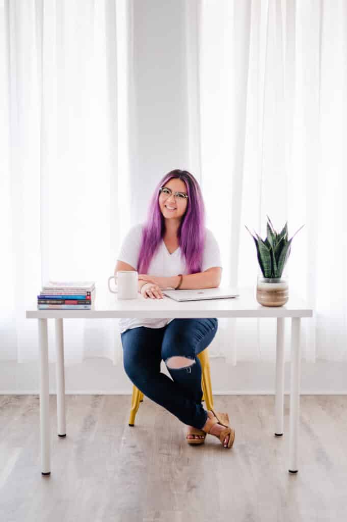 Smiling woman with purple hair at a bright, modern desk filled with books.