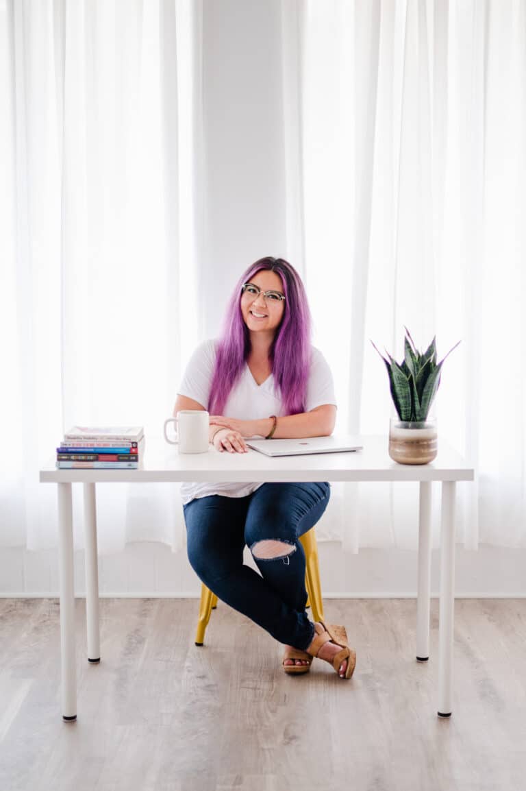 Smiling woman with purple hair at a bright, modern desk filled with books.
