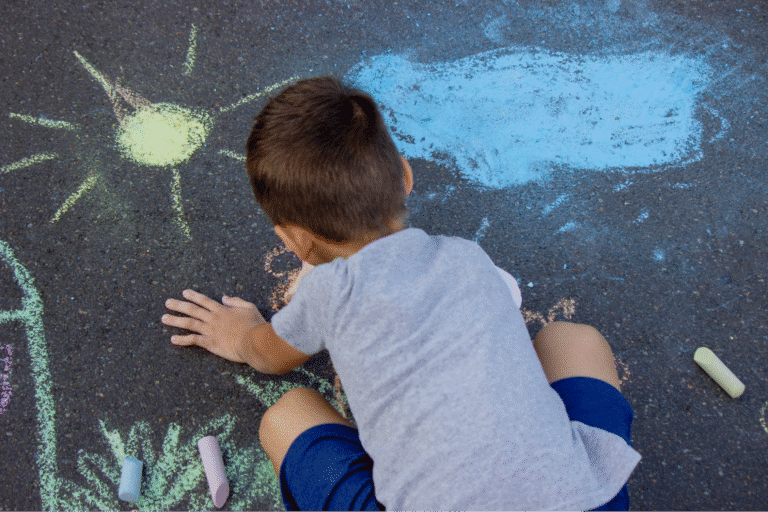 Child joyfully drawing with colorful chalk on pavement under the sun.