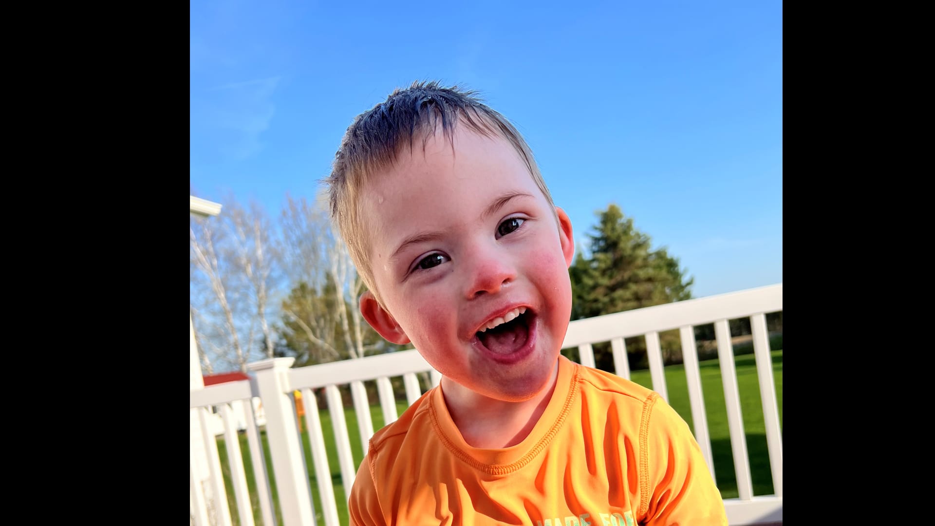 Joyful child smiling outdoors in a vibrant orange shirt under a clear blue sky.