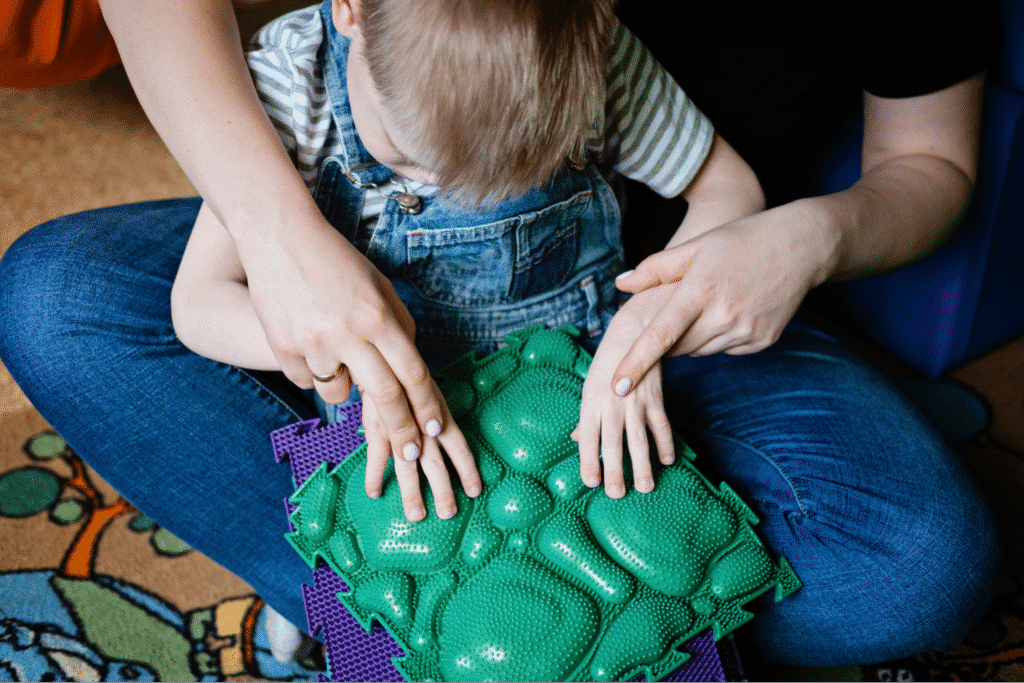 Child engaging in sensory play with tactile green mats and adult assistance.