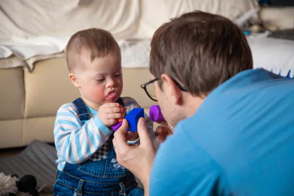 A joyful baby boy with Down syndrome plays with colorful toys alongside his dad.