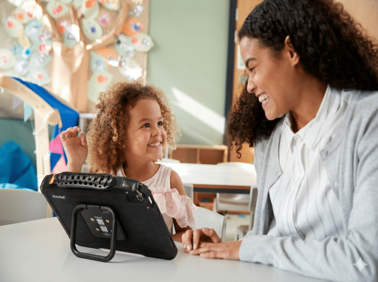 Happy SLP engaging with child while using a Quicktalker Freestyle AAC device in a bright classroom setting.
