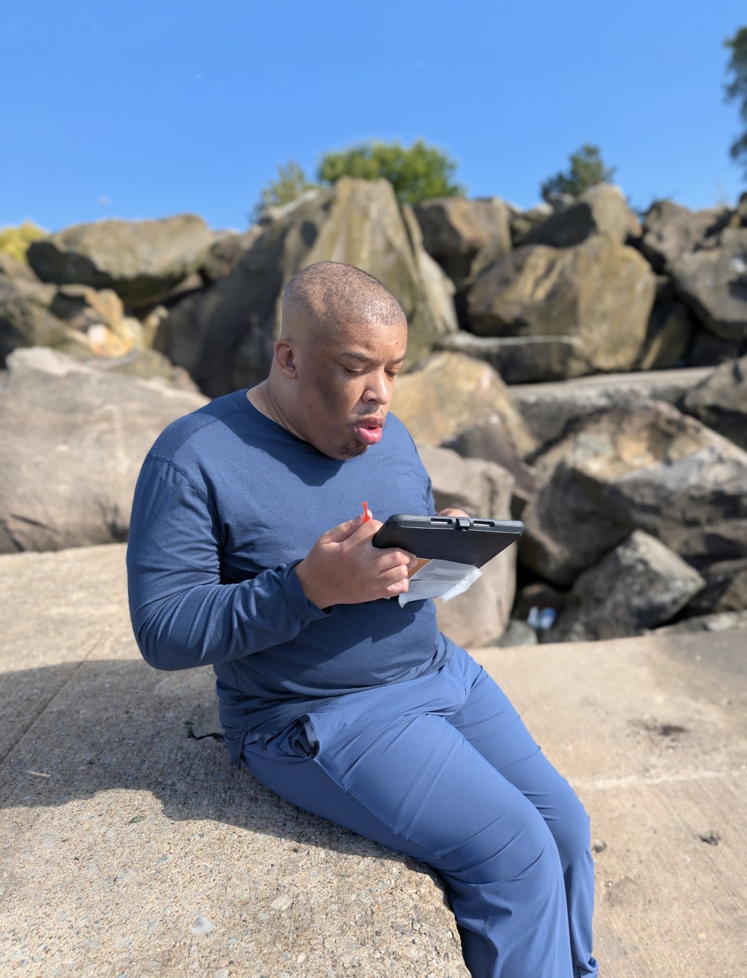Man reading on a tablet by the rocks under a clear blue sky.