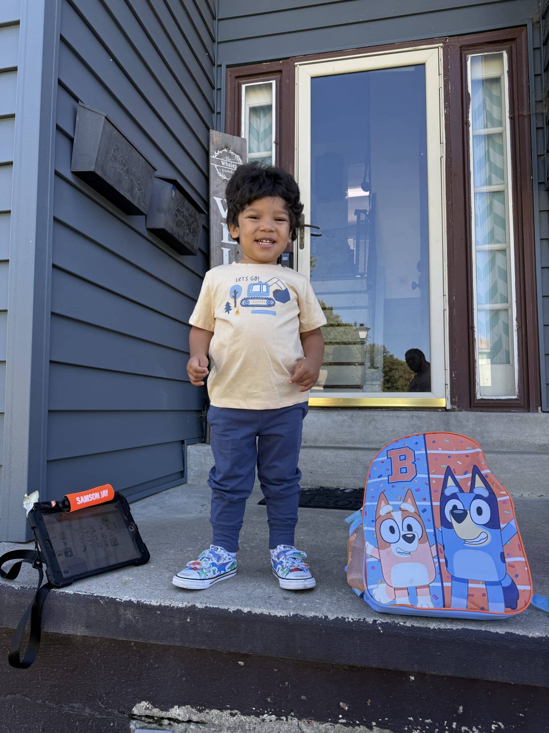 Happy little boy standing on porch with backpack and tablet, ready for adventure.