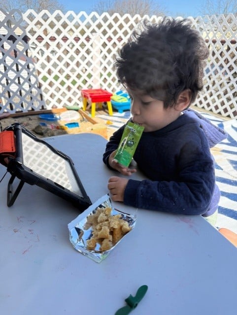 Child enjoying snack and screen time outdoors during playtime.