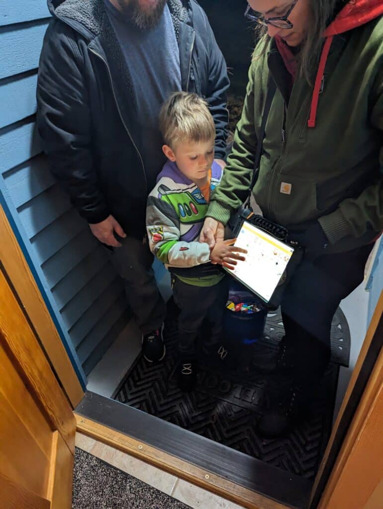 Logan, dressed in a costume, uses his QuickTalker Freestyle at a front door while two adults look on.