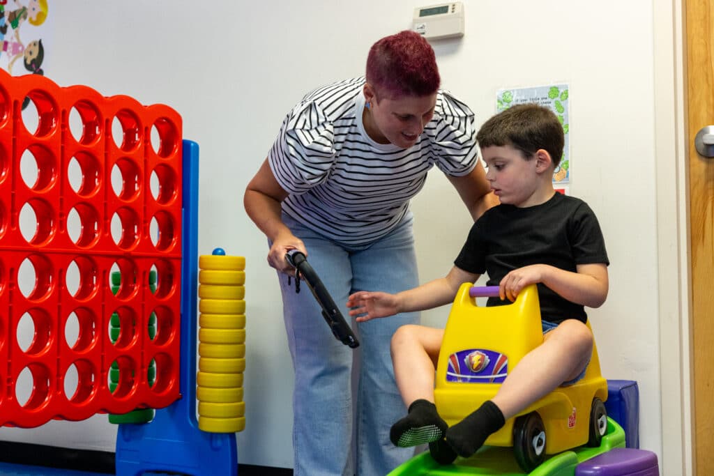 A speech-language pathologist holds a QuickTalker Freestyle speech device while a boy plays in a therapy room