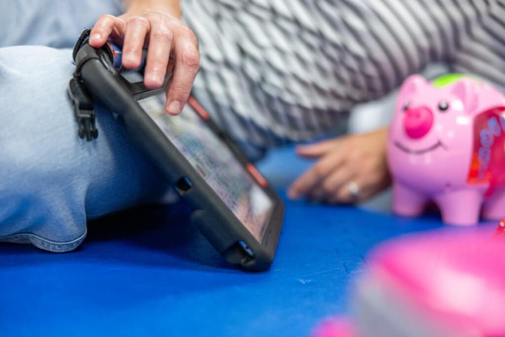 A close view of hands using a QuickTalker Freestyle speech device on a floor mat during a play-based therapy session