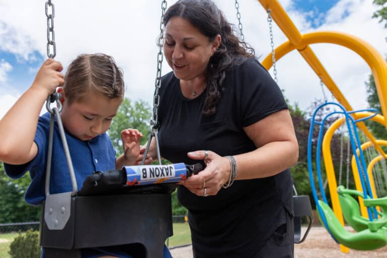 A child plays on a swing while an adult assists with a Quicktalker Freestyle at the park.