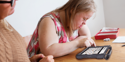 Two women enjoy playing bingo on a high-tech AAC device at a wooden table.