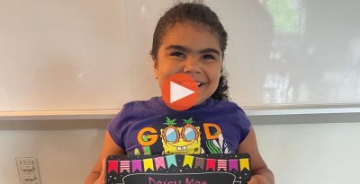 Daisy Mae smiles proudly holding her name sign in a colorful classroom setting.