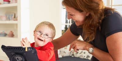 Child joyfully learning with a tablet alongside a smiling caregiver in a bright setting.