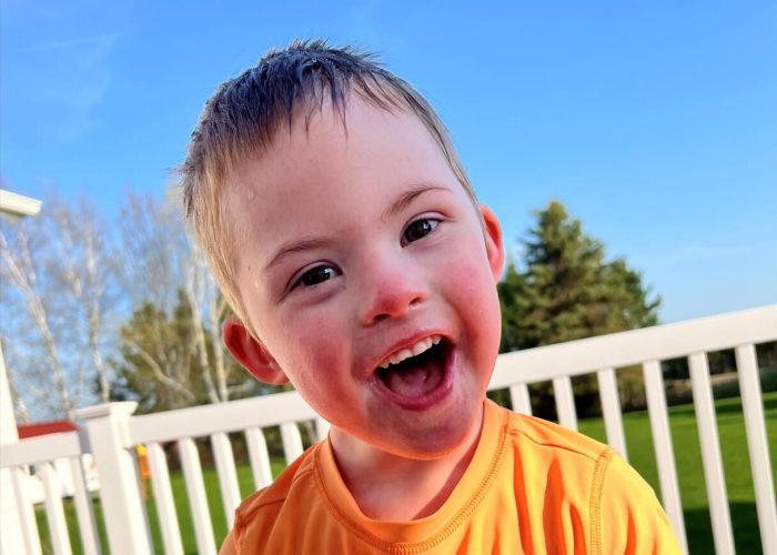 A joyful child in an orange shirt smiling outdoors on a sunny day.