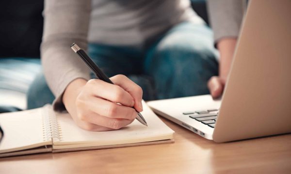 Woman taking notes while studying online with a laptop at her work desk.